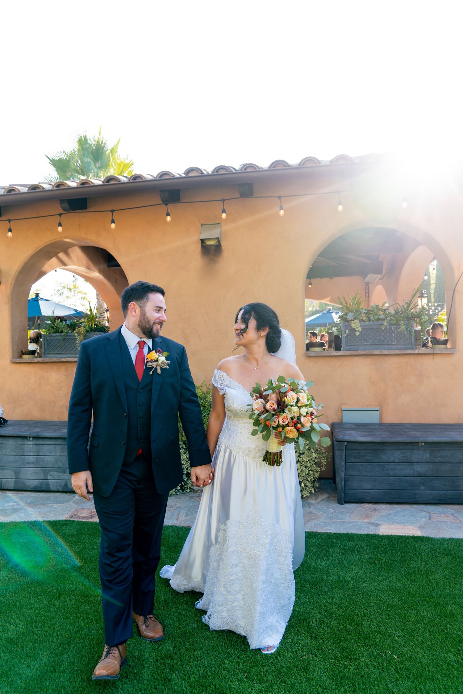 Newlyweds walk together on artificial turf in front of a Spanish-style venue at sunset.