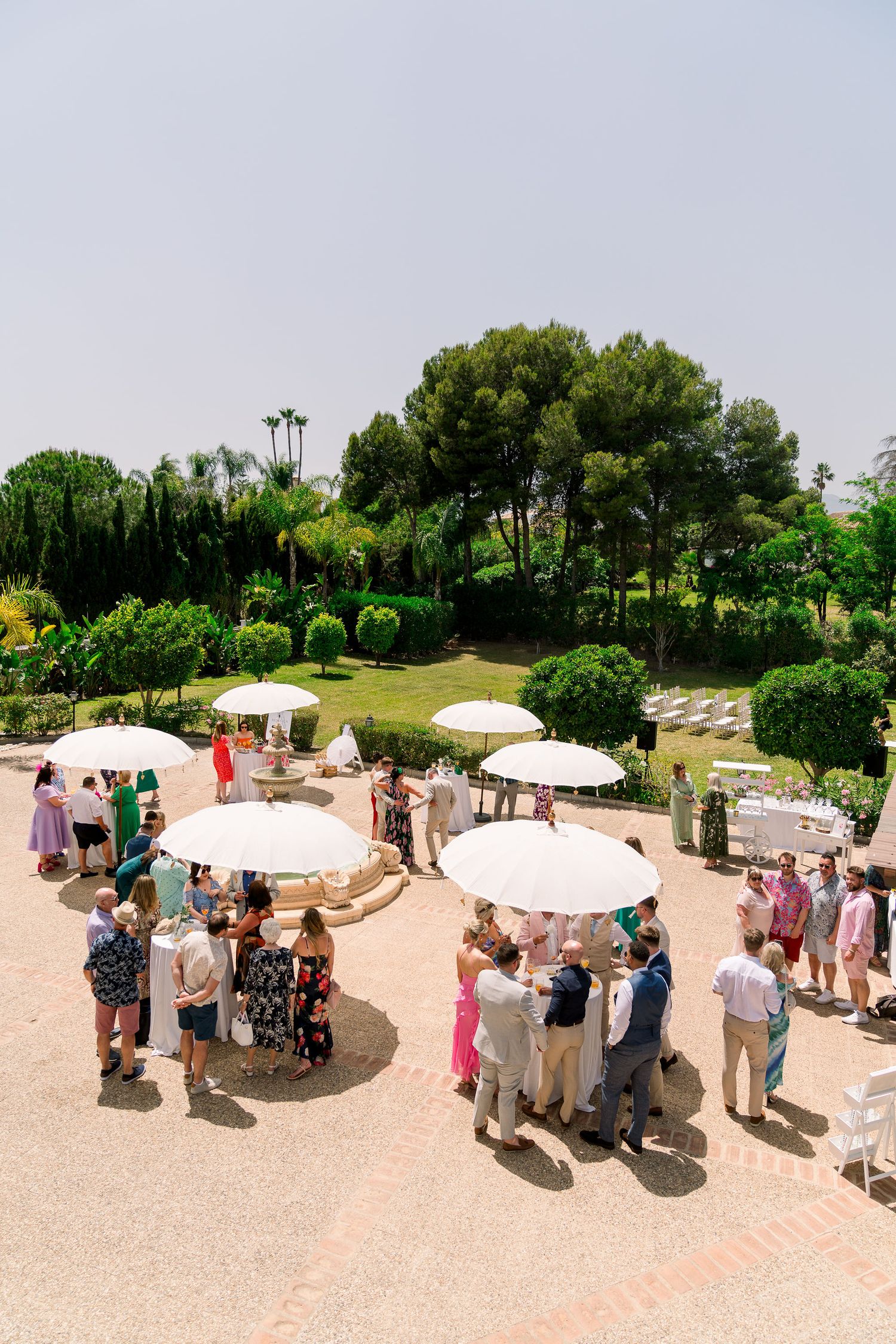 Guests gather under white umbrellas during an outdoor garden party with lush greenery and palm trees in the background.