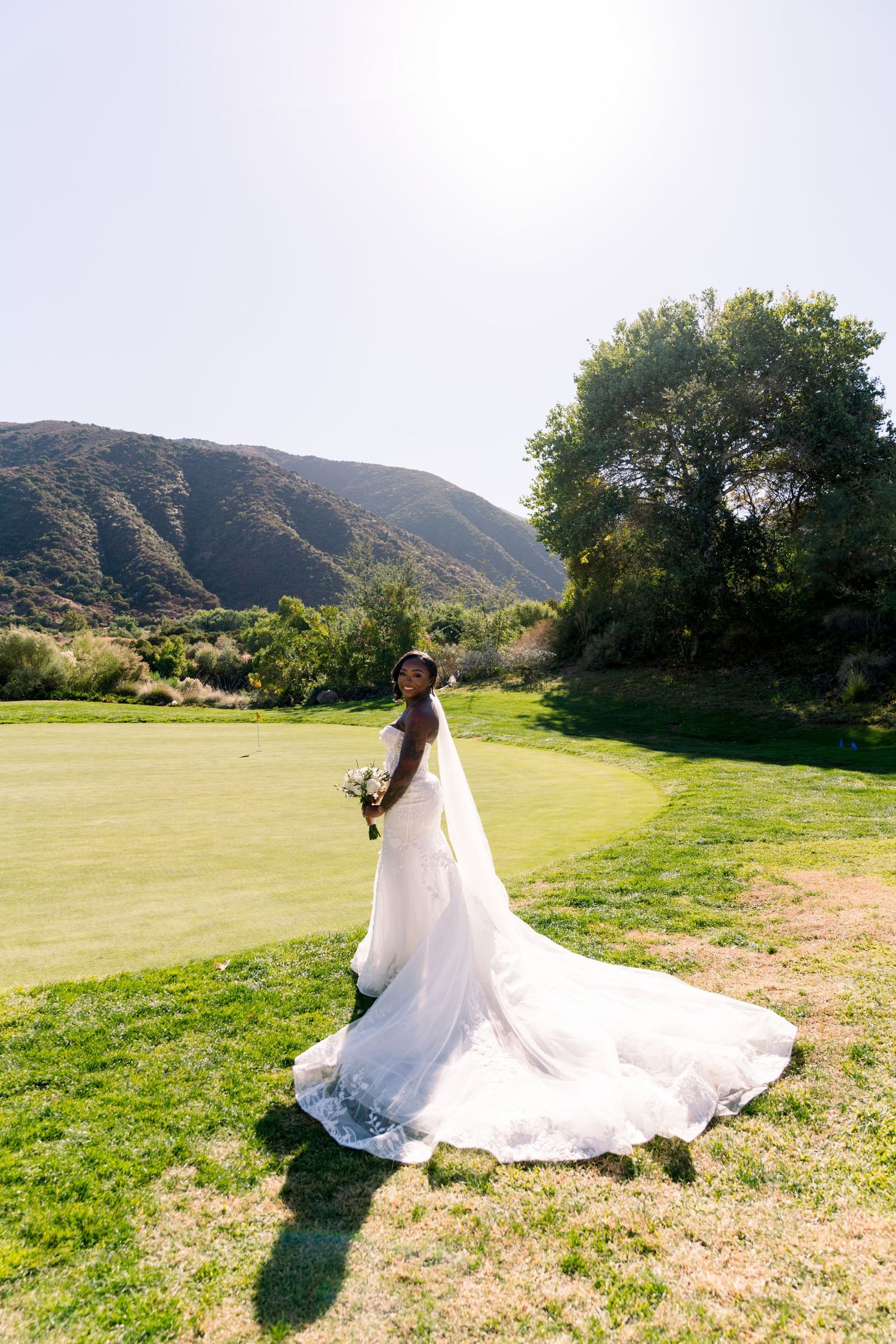 A flowing white wedding dress with long train photographed against mountain landscape on green grass.