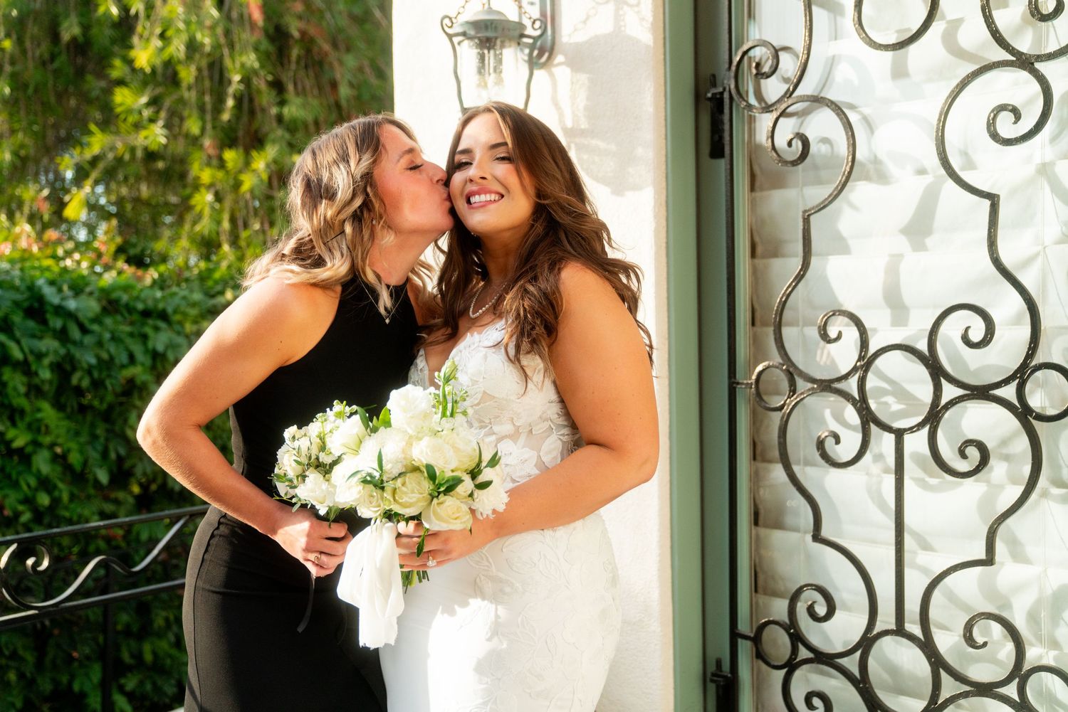 Two women share a joyful moment with white bouquet while posing by an ornate door.