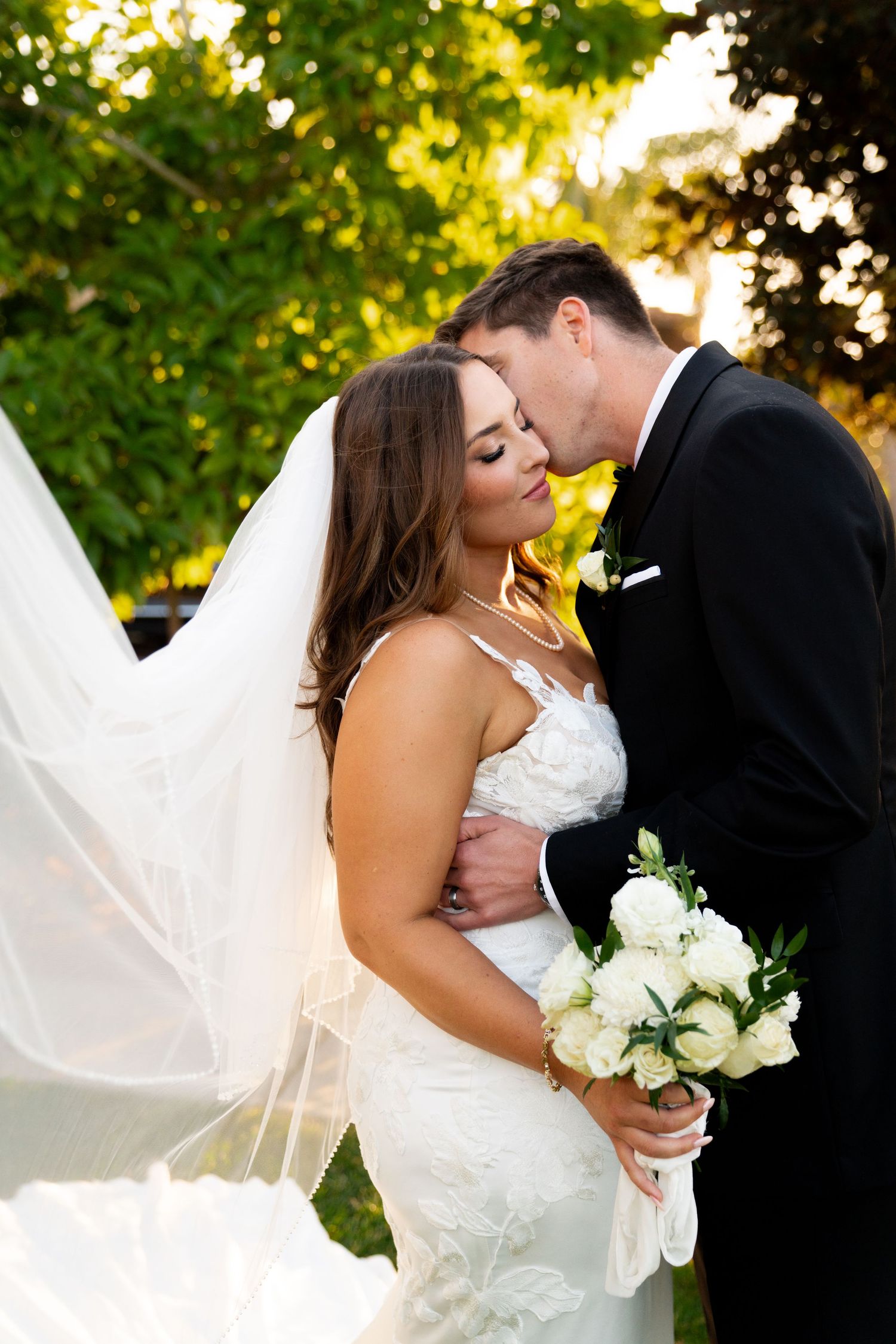 A romantic sunset wedding photo featuring a couple sharing an intimate moment with a flowing veil and white bouquet.