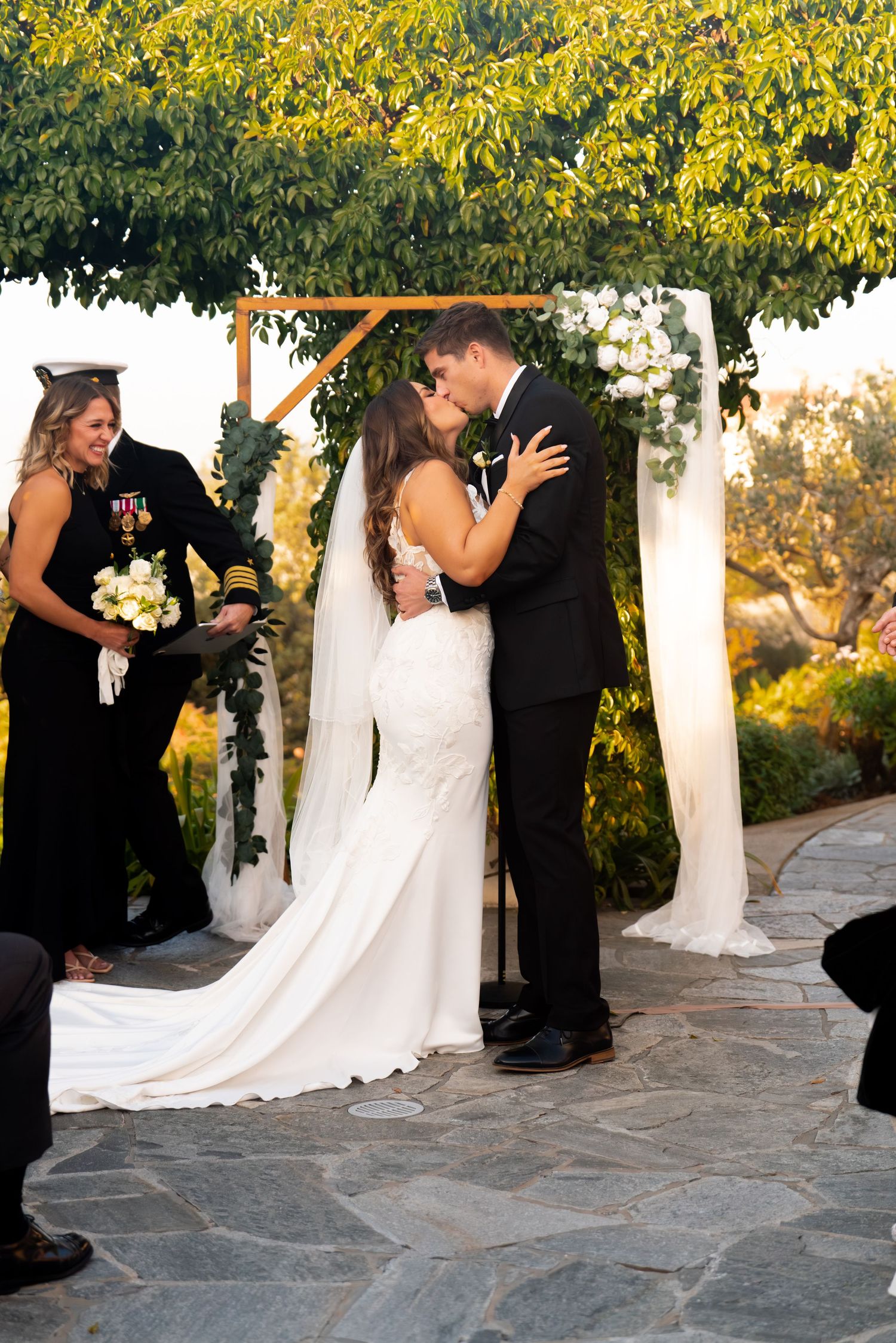 Newlyweds share their first kiss under a tree arch during an outdoor wedding ceremony.
