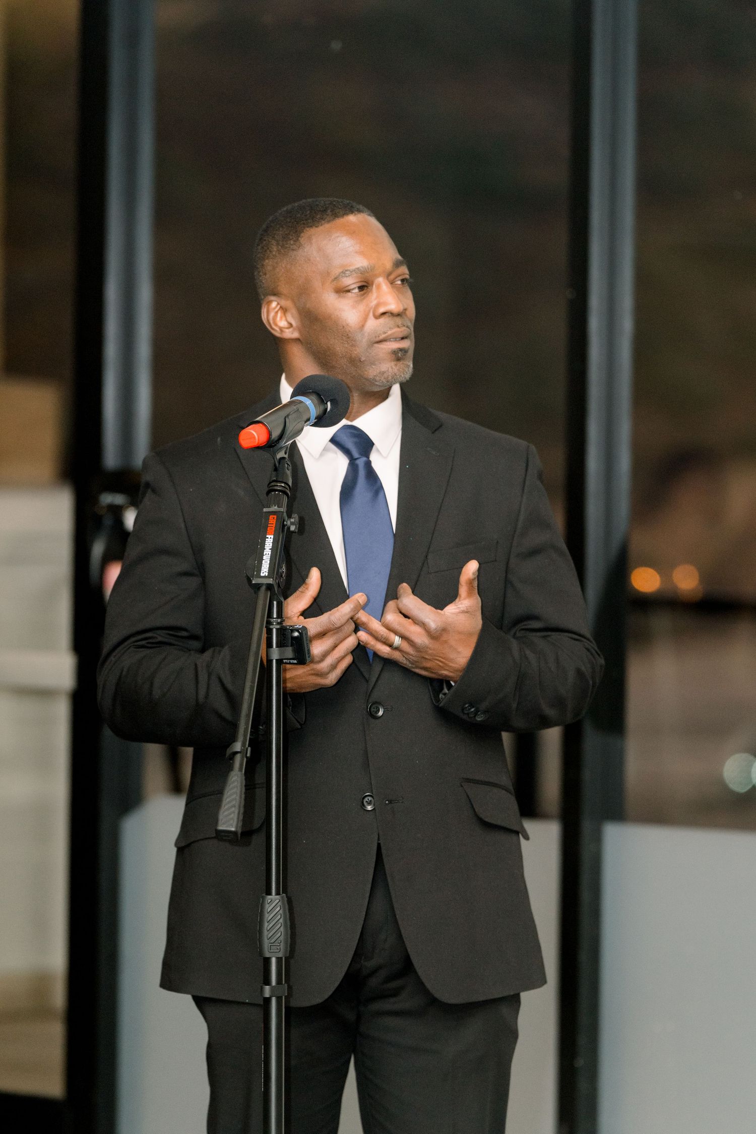 Speaker in dark suit delivers speech at formal event with microphone.