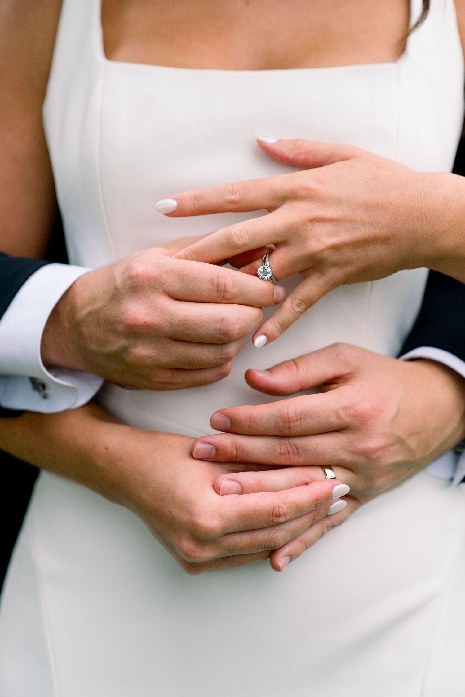 Close-up of hands embracing while showing an engagement ring on a white dress background.