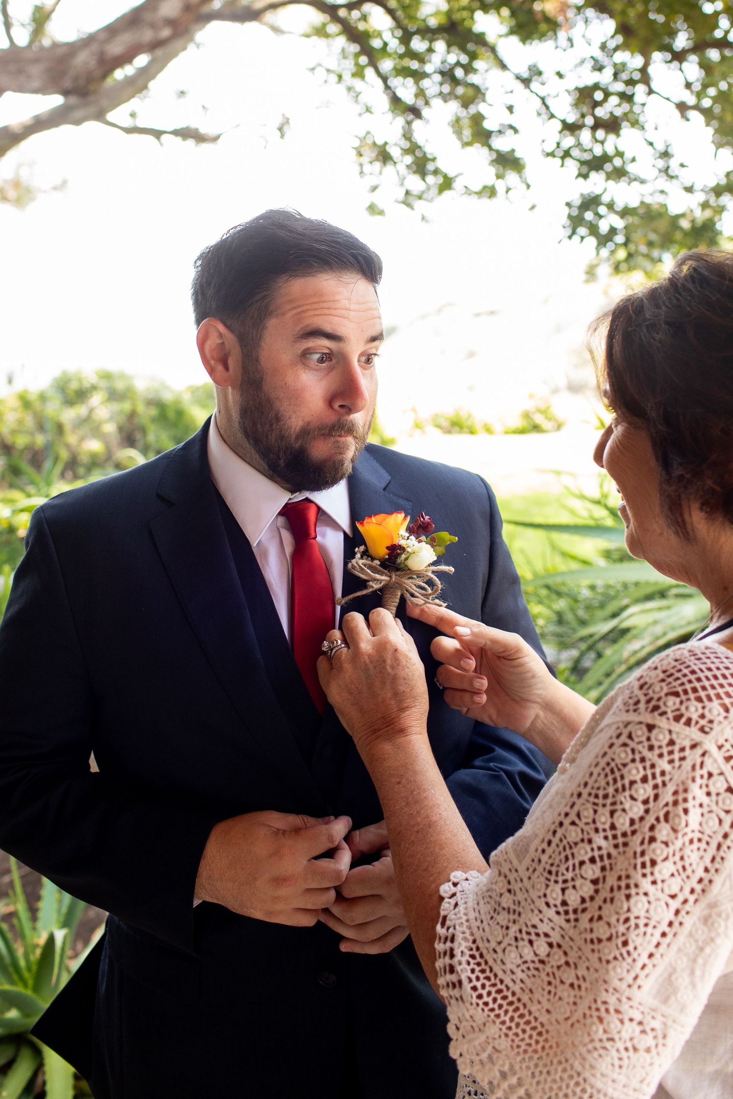 A boutonniere is being pinned onto a navy suit jacket with a red tie during a wedding preparation moment.