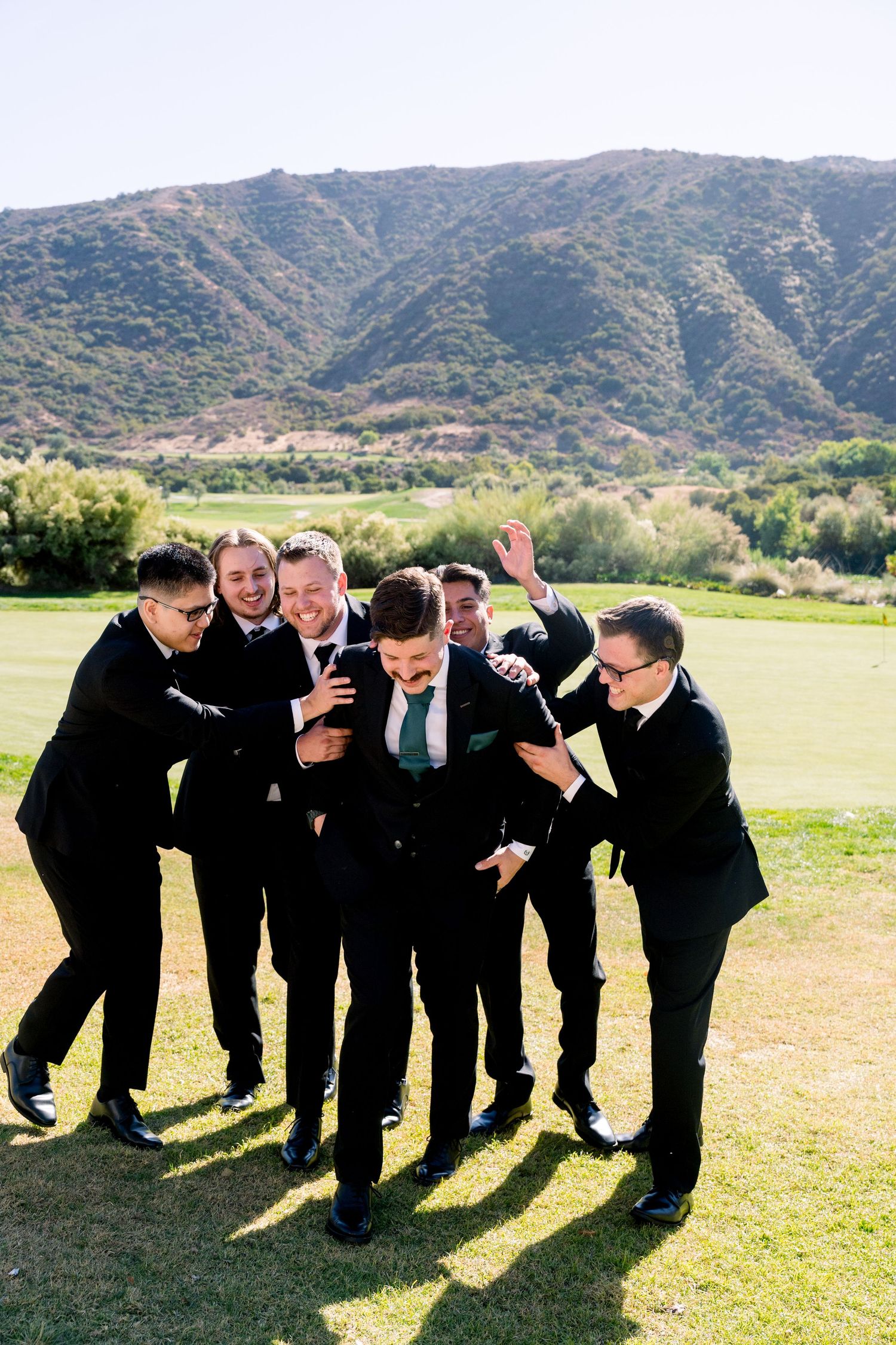A group of groomsmen in black suits playfully huddle together on a lawn with mountains in the background.