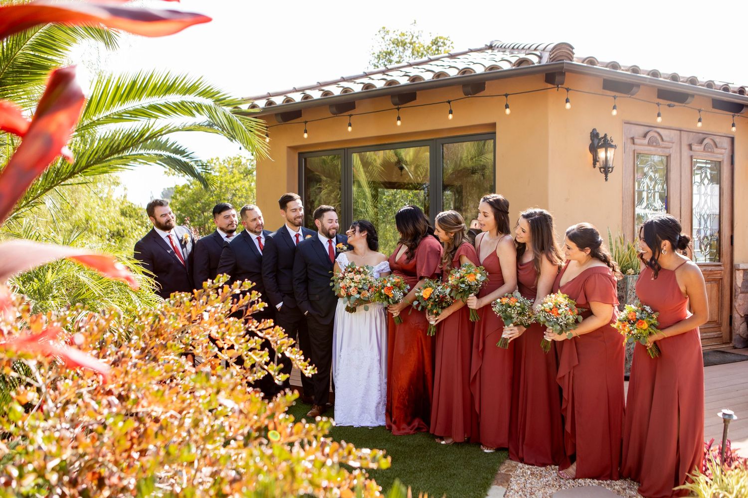 Wedding party in navy suits and burgundy dresses posed together outside Spanish-style venue.