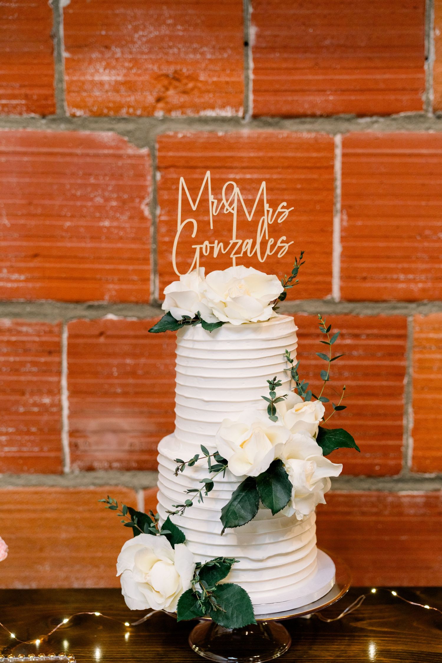 Elegant three-tiered white wedding cake with fresh roses and greenery against red brick wall, featuring Mr Mrs Gonzalez topper.