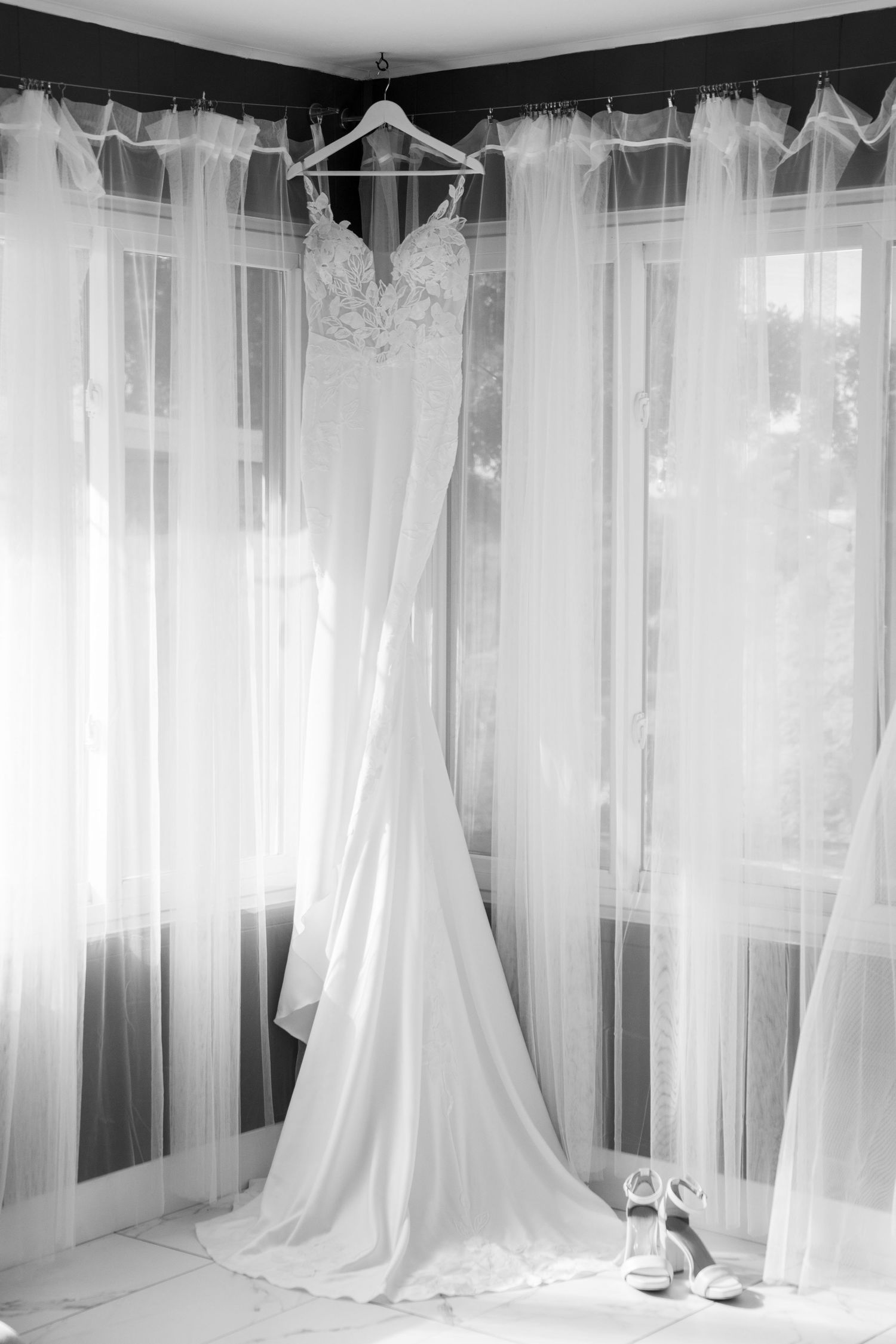 An elegant white lace wedding dress with train hangs in front of sheer curtains with bridal shoes below.