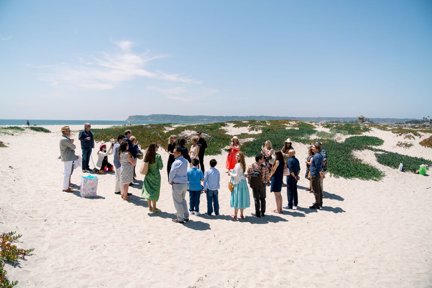 A group of people standing in a circle on a sandy beach with dunes and blue sky in the background.