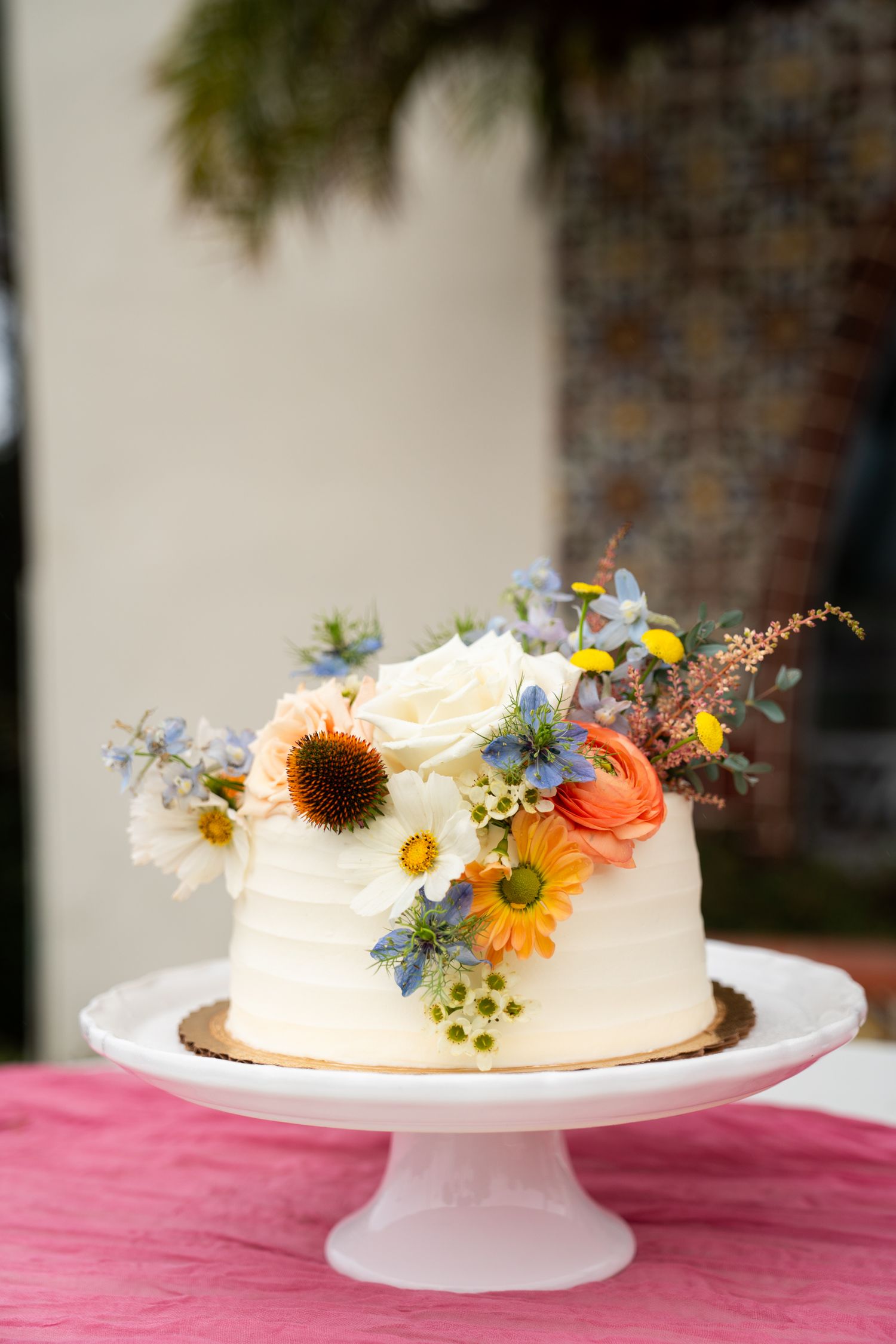 A rustic single-tier white wedding cake decorated with wildflowers including sunflowers and daisies on a pink tablecloth.
