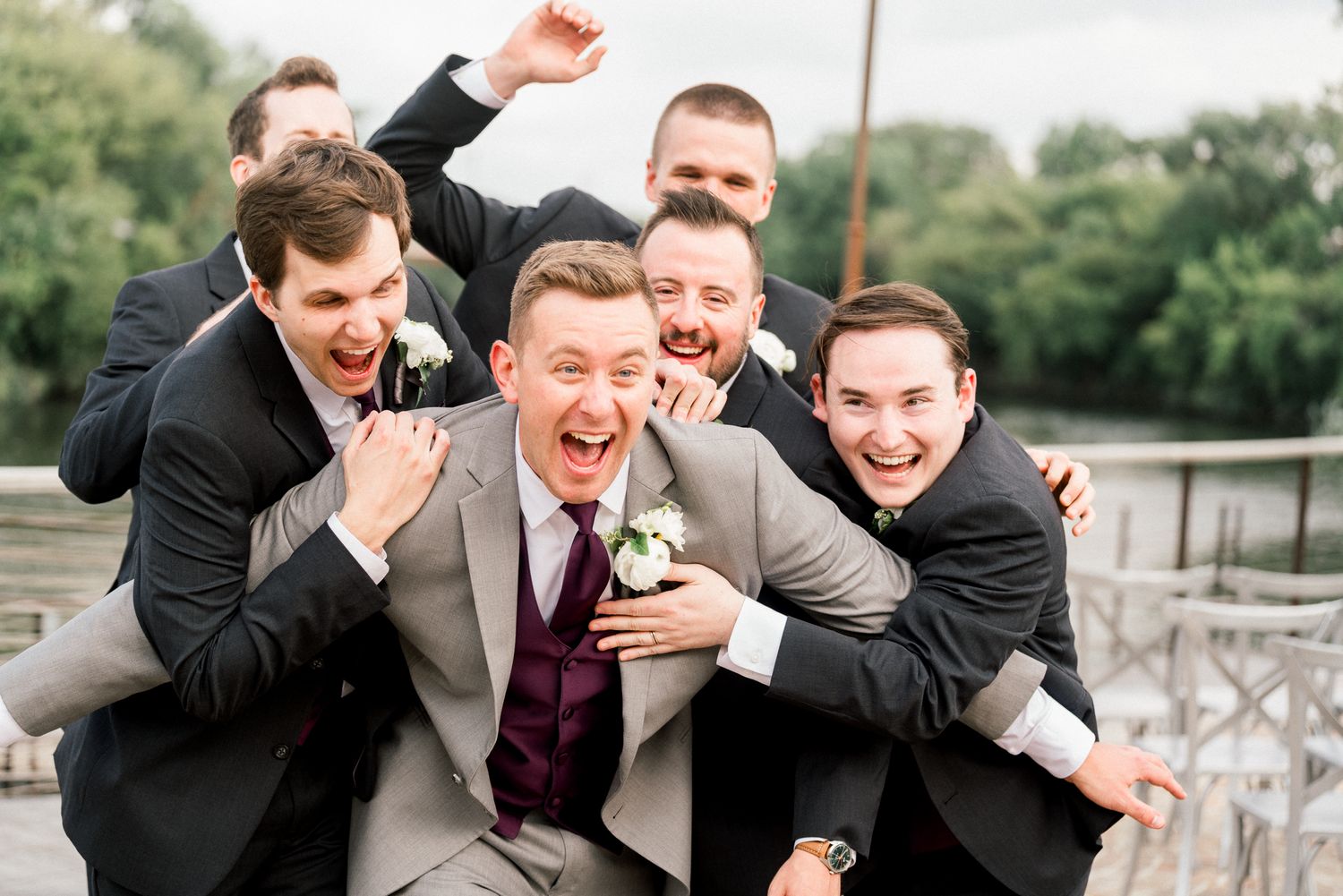 A group in matching black suits and purple tie pose playfully together for a candid wedding party photo by the lake.