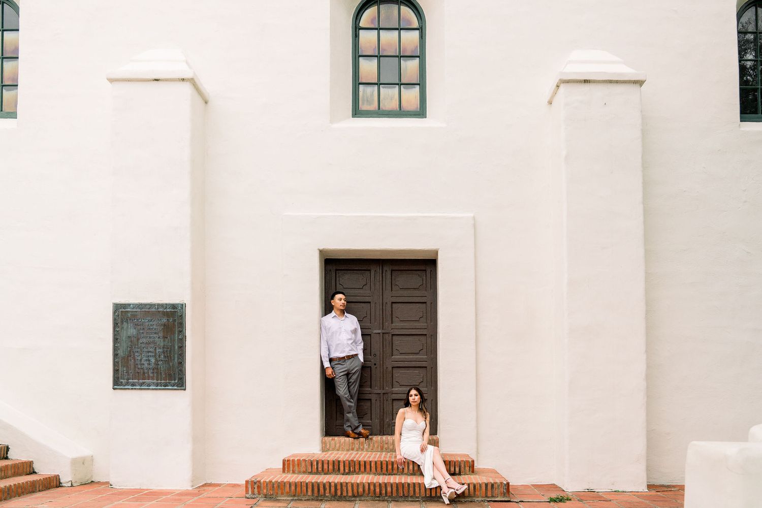 A figure stands with their dog on brick steps in front of a dark wooden door on a white mission-style building.