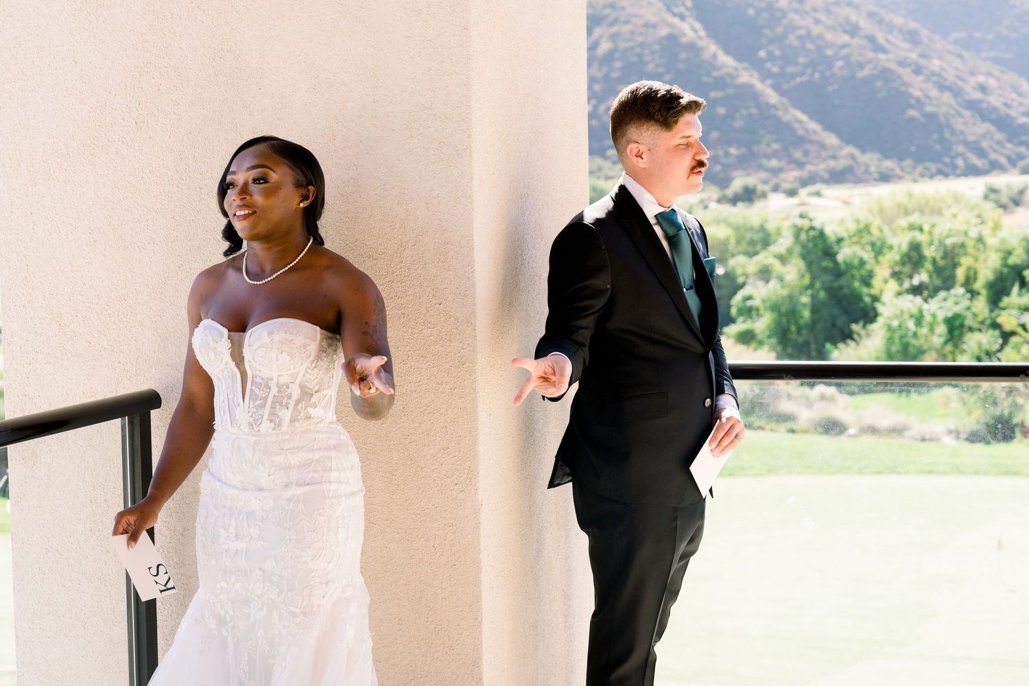 Couple in wedding attire on opposite sides of a wall for a first look moment with mountains in background.
