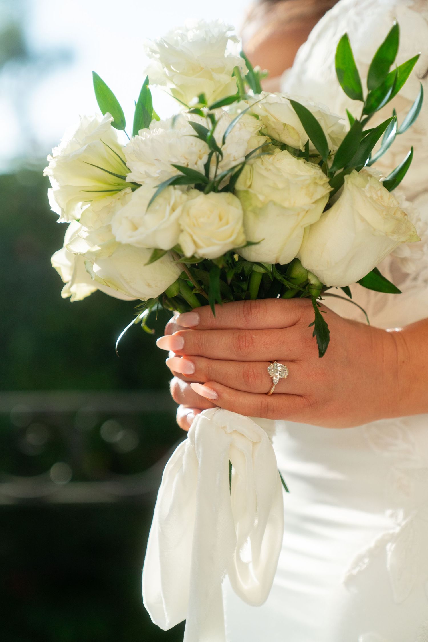White rose wedding bouquet held by hands with an engagement ring against a light background.