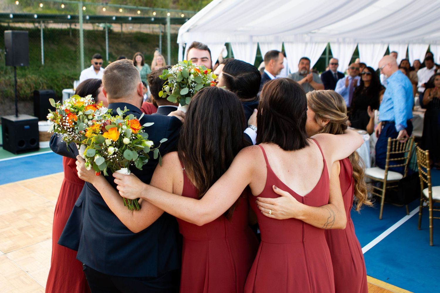 Bridesmaids in burgundy dresses embrace while holding yellow sunflower bouquets at outdoor reception.