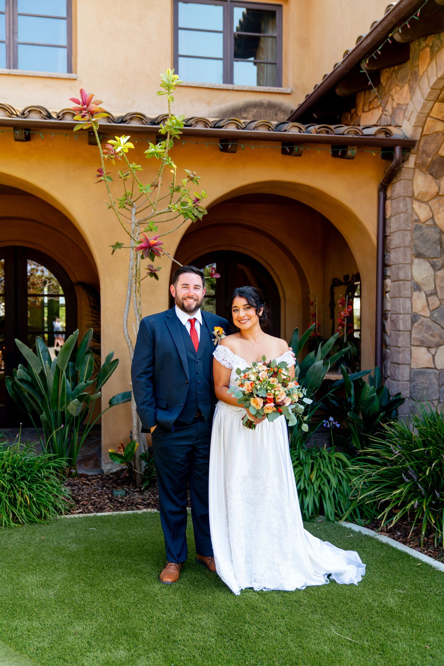 A wedding portrait captured in front of a Spanish-style building with stone archways and tropical landscaping.