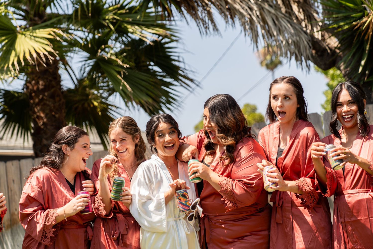 A group in matching coral-colored satin robes share drinks and laughter under palm trees at an outdoor celebration.