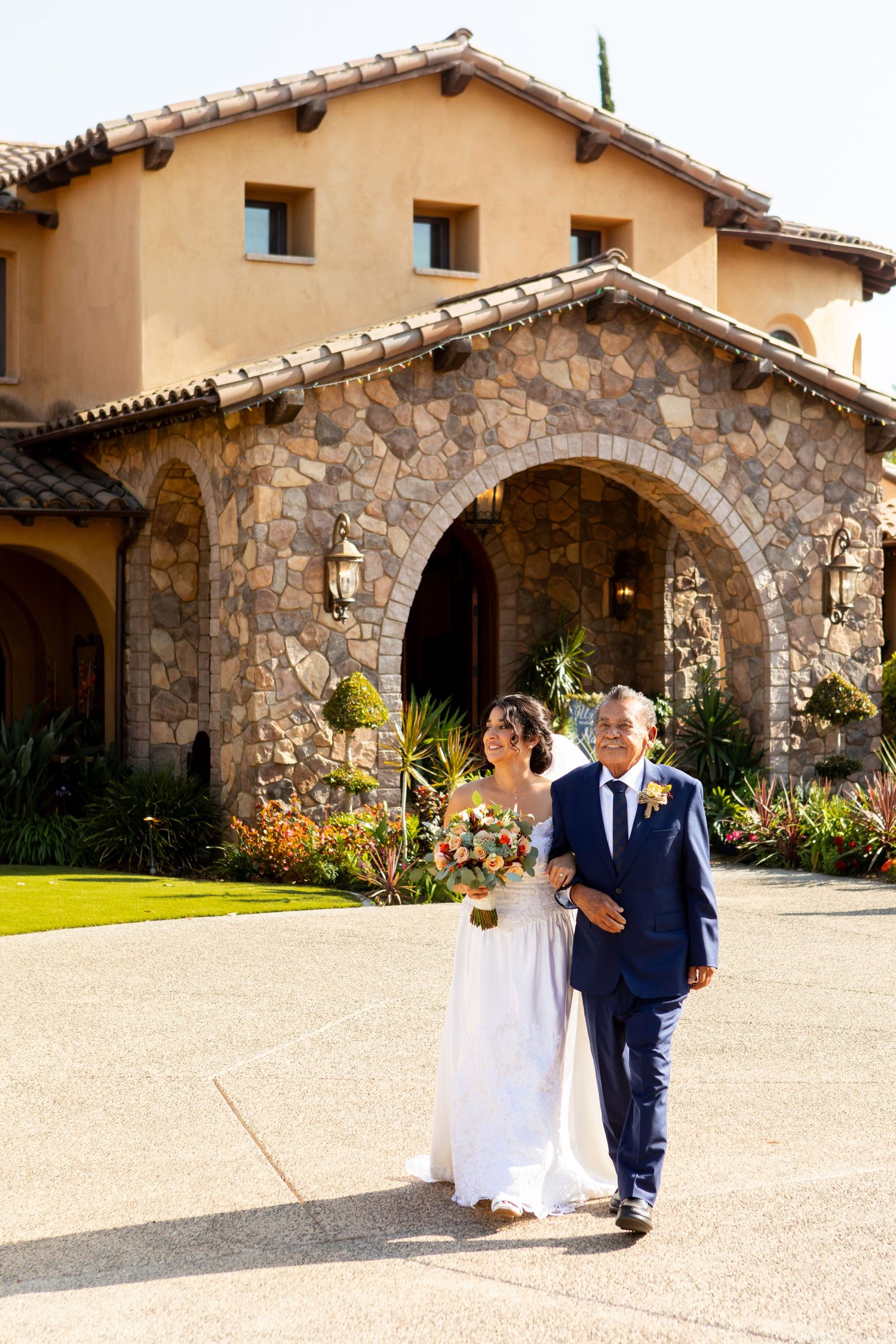 A couple walks together outside a Mediterranean-style stone building with arched entryway and terra cotta roof tiles.