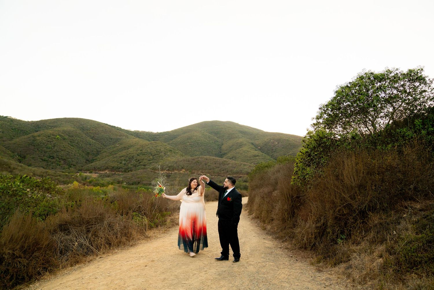Two people dance together on a scenic dirt path surrounded by rolling green hills and brush.