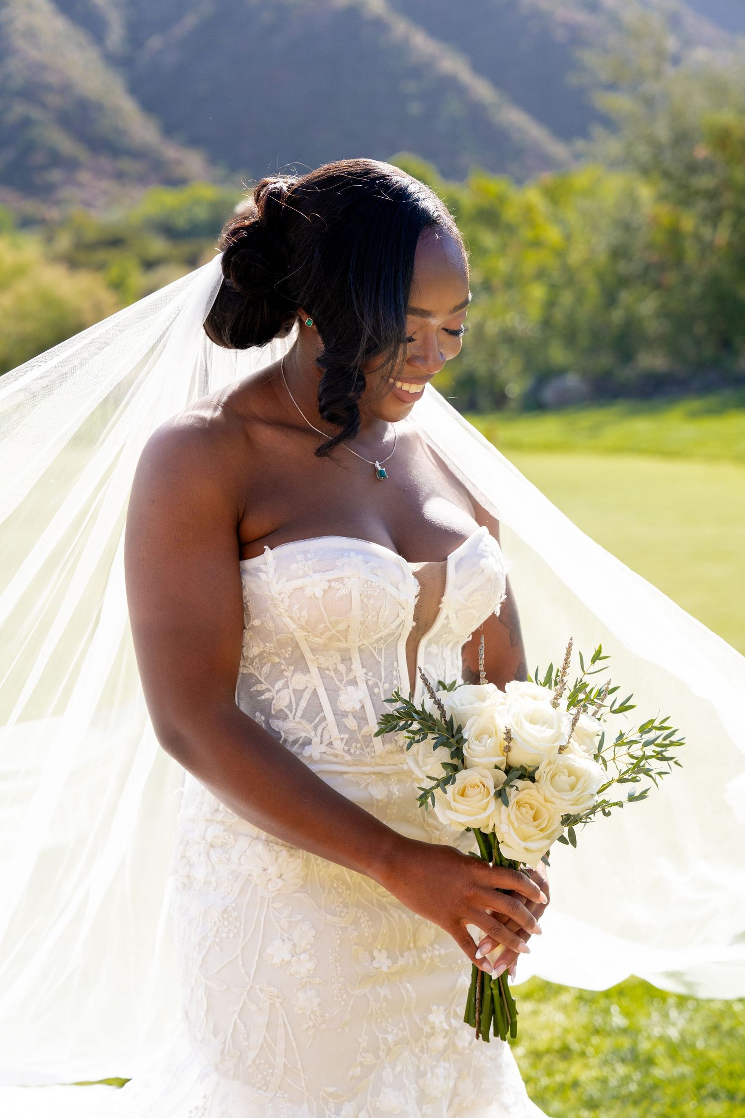 Bride in white lace wedding dress holding bouquet of white flowers against scenic mountain backdrop.