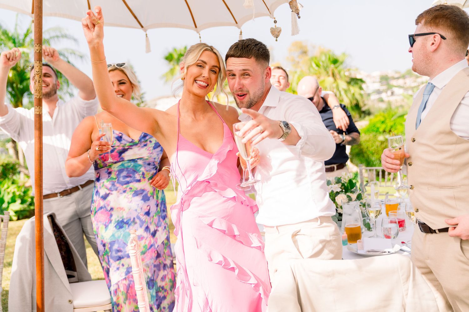 Joyful celebration under a white tent with wedding guests in floral and pink dresses raising their hands in celebration.