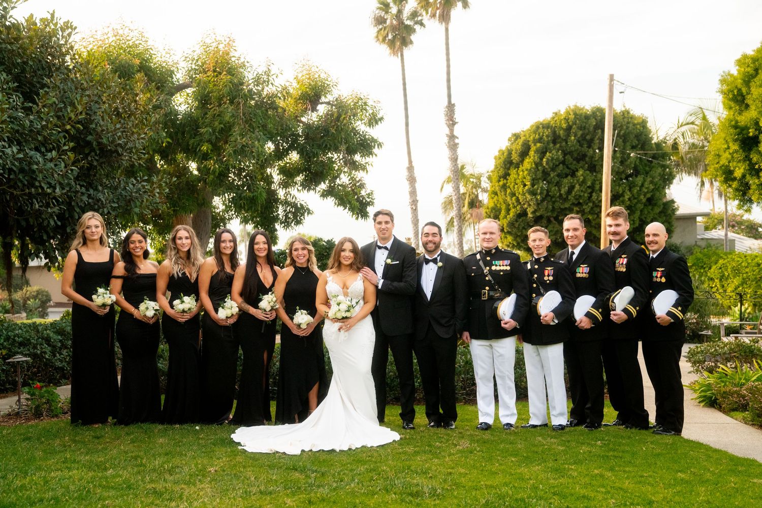 Wedding party in formal black attire and white uniforms poses together on a garden lawn with palm trees.