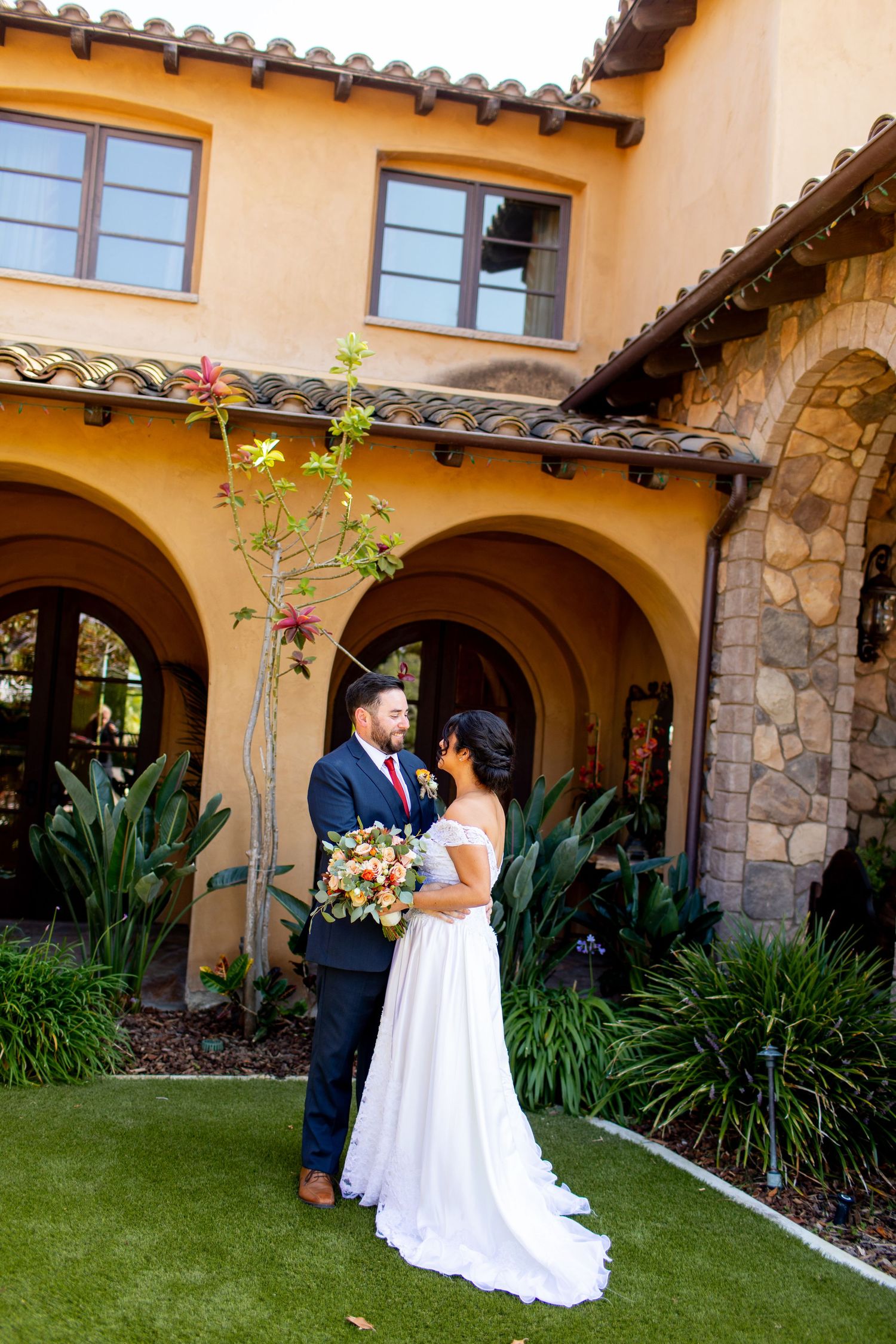 A wedding couple stands together under Mediterranean-style arches surrounded by lush landscaping and Spanish tile roof details.