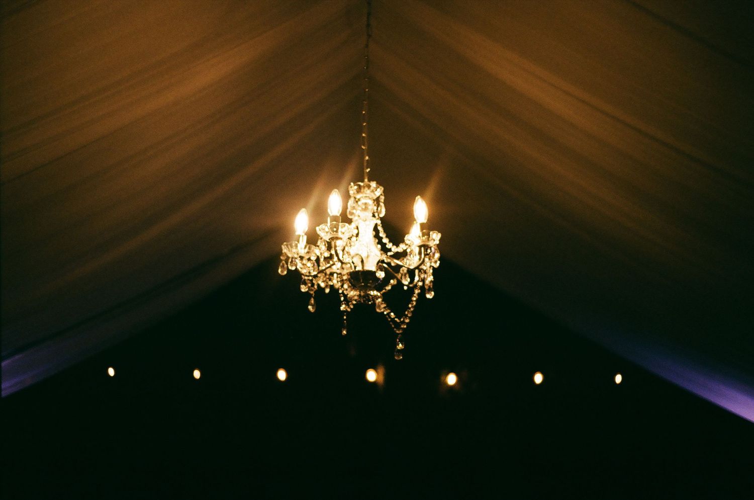 Elegant crystal chandelier hanging in dark tent ceiling with string lights creating ambient glow.