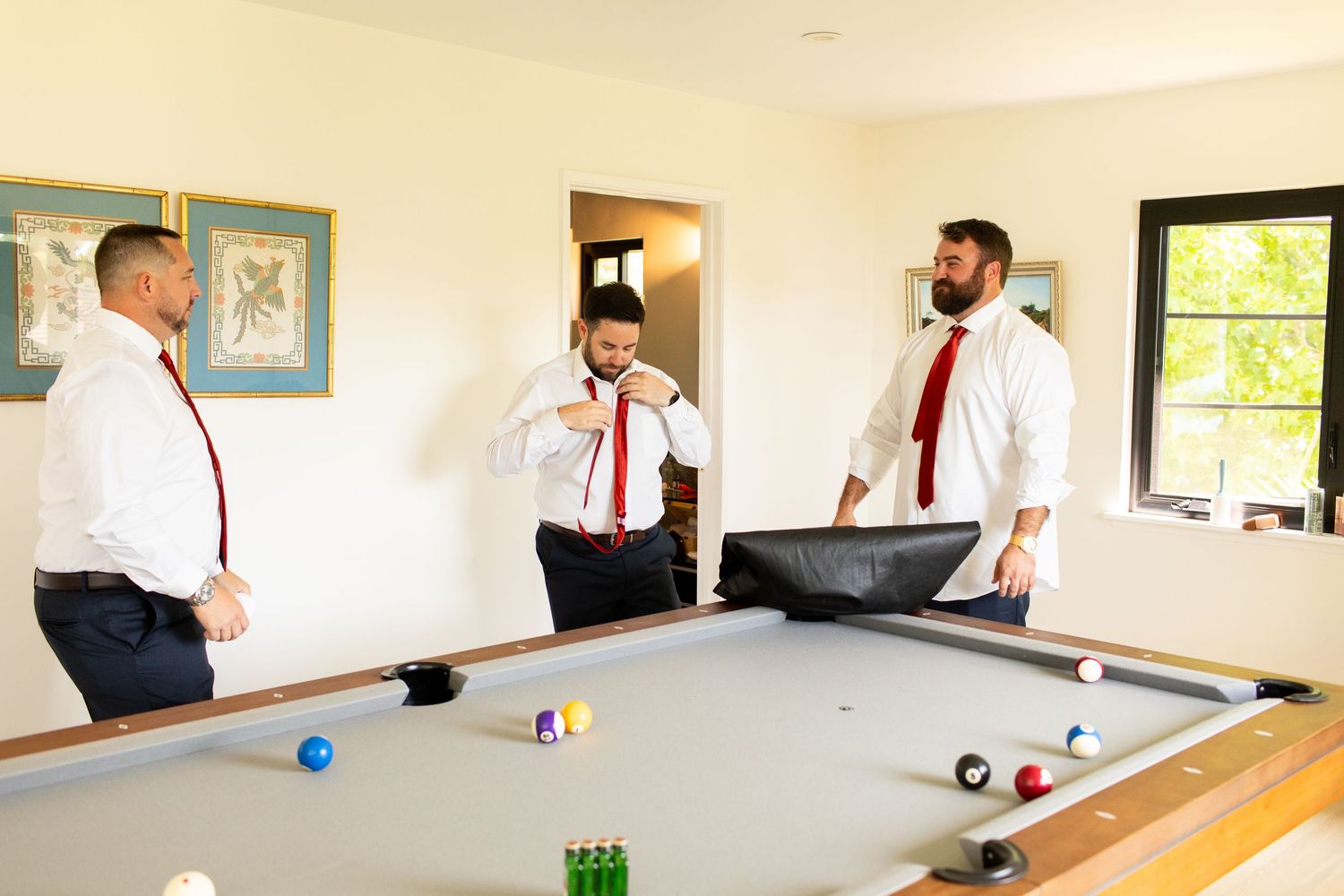 Three people in white dress shirts and red ties play pool together in a bright room with framed artwork.