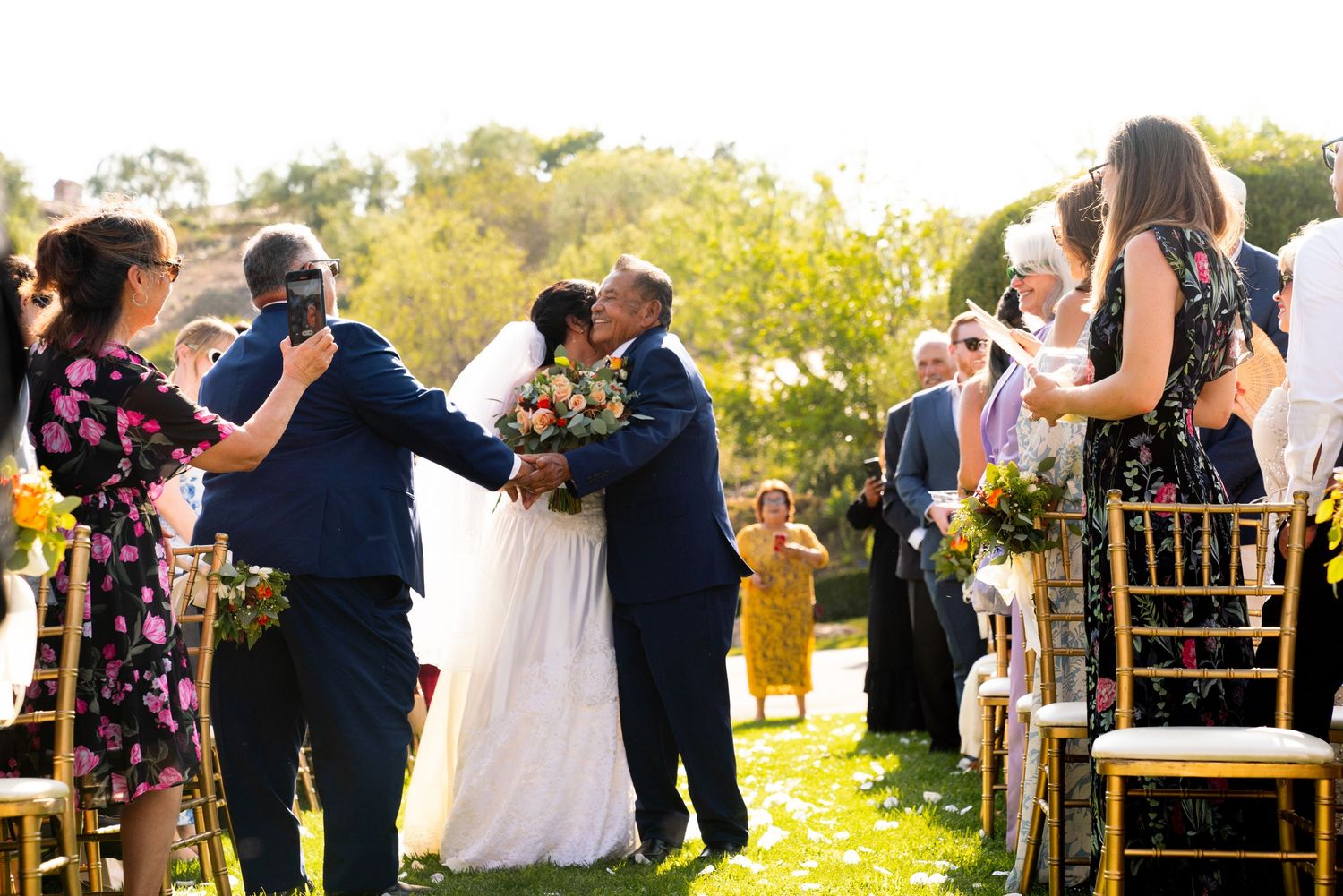 An outdoor wedding ceremony takes place in a garden setting with guests seated on gold chairs during golden hour.