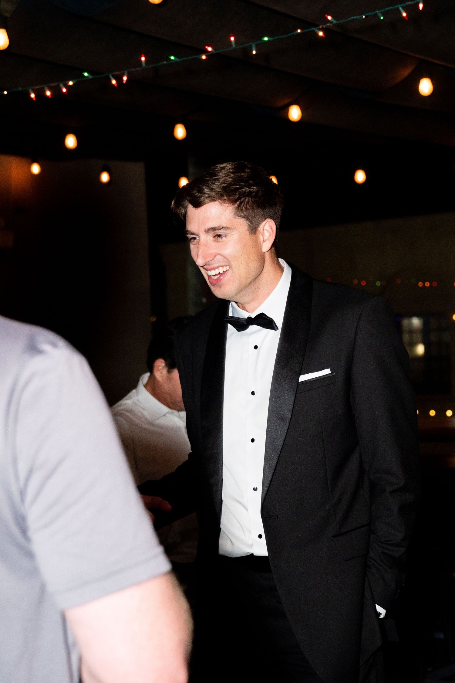 Guest in formal black tuxedo and bowtie enjoying conversation at dimly lit reception.