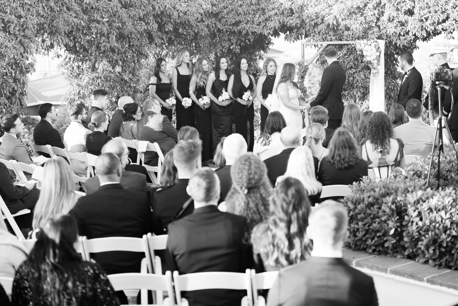 A group of performers in formal attire stand in rows during an outdoor ceremony with seated audience members.