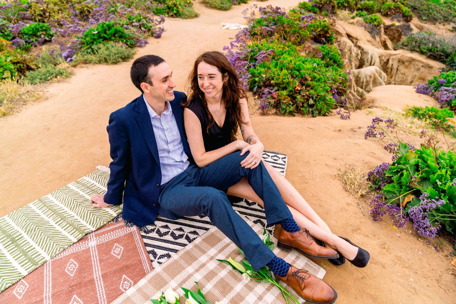 Two people sit together on a patterned blanket along a garden path surrounded by colorful flowers and coastal plants.
