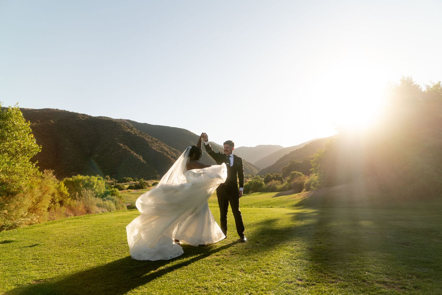 A wedding couple stands in a sunlit field with mountains and dramatic lighting in the background.