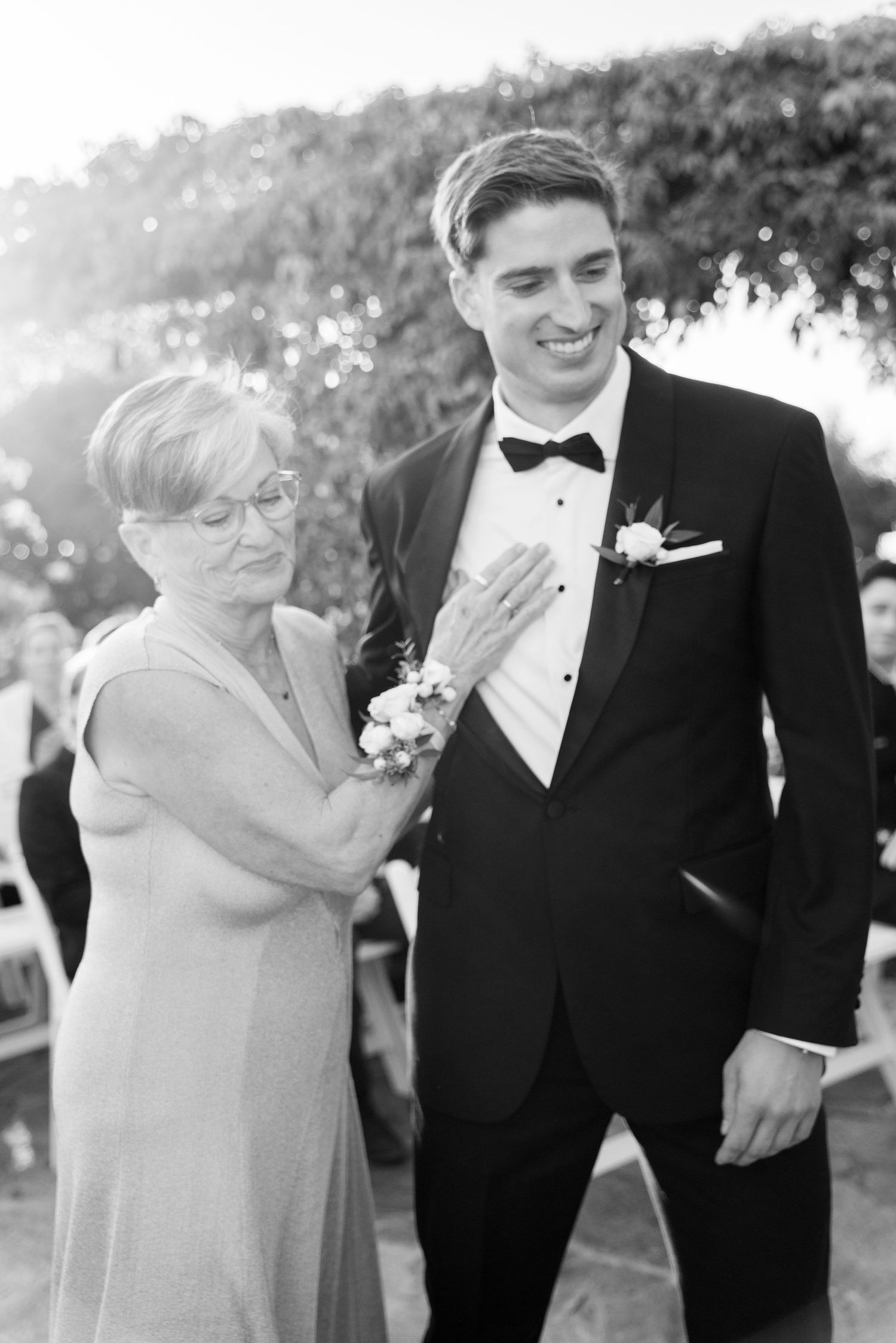 A black and white photo of a tender moment between a son in a tuxedo and his mother at a wedding.