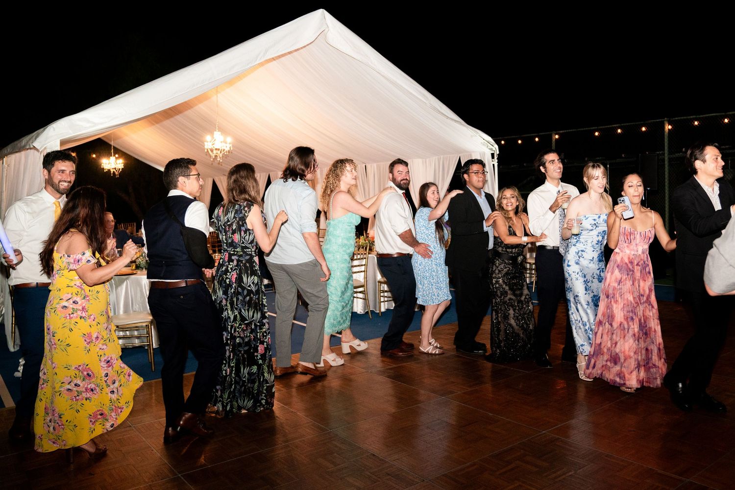 Party guests dance and celebrate on a wooden dance floor under a white tent at an evening event.