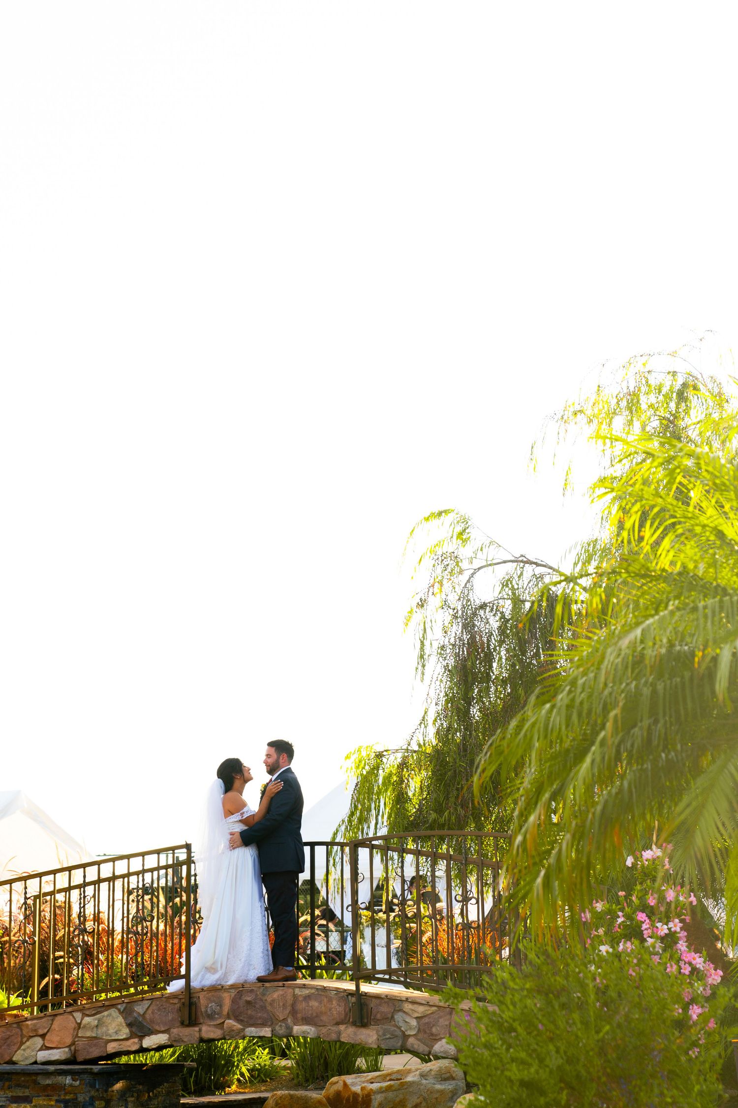 Wedding couple stands on stone bridge with weeping willow tree in romantic garden setting.