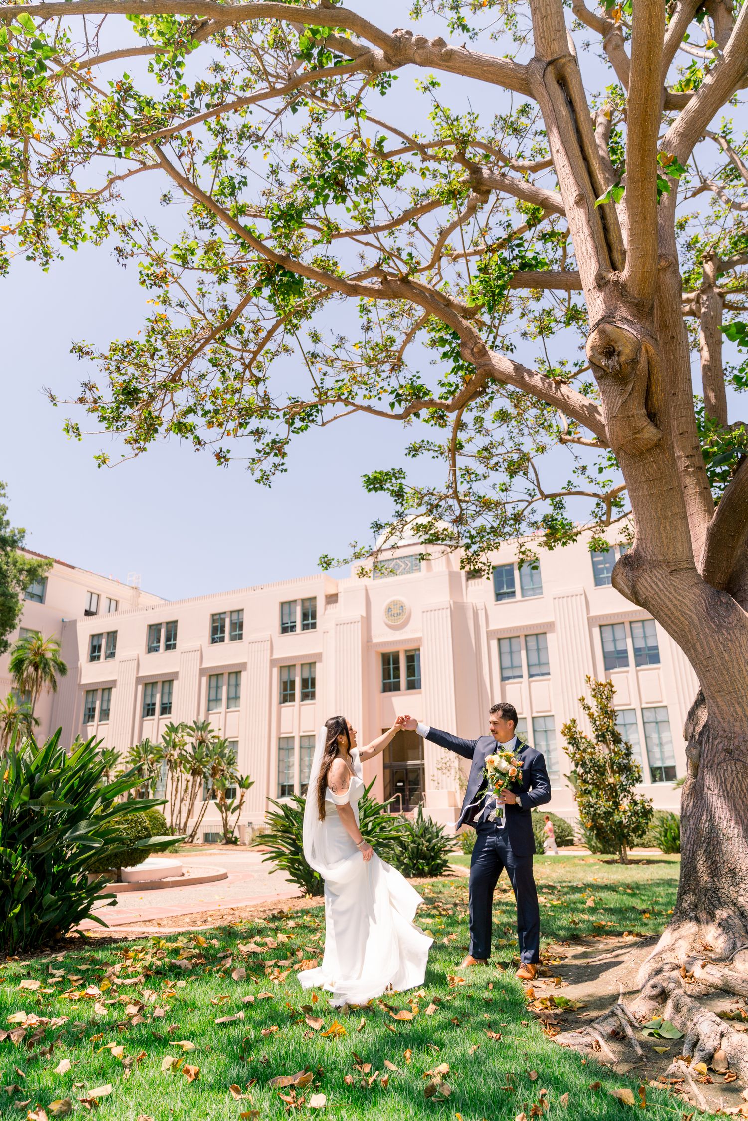 Happy couple stands together under large shade tree with pink building in background on sunny wedding day.
