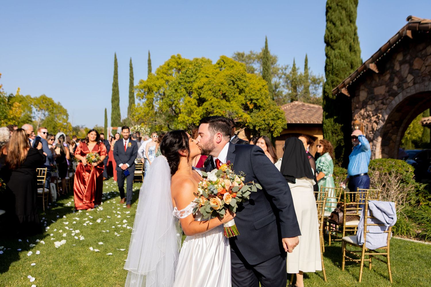 A joyful outdoor wedding ceremony with flower petals scattered on the grass under cypress trees.