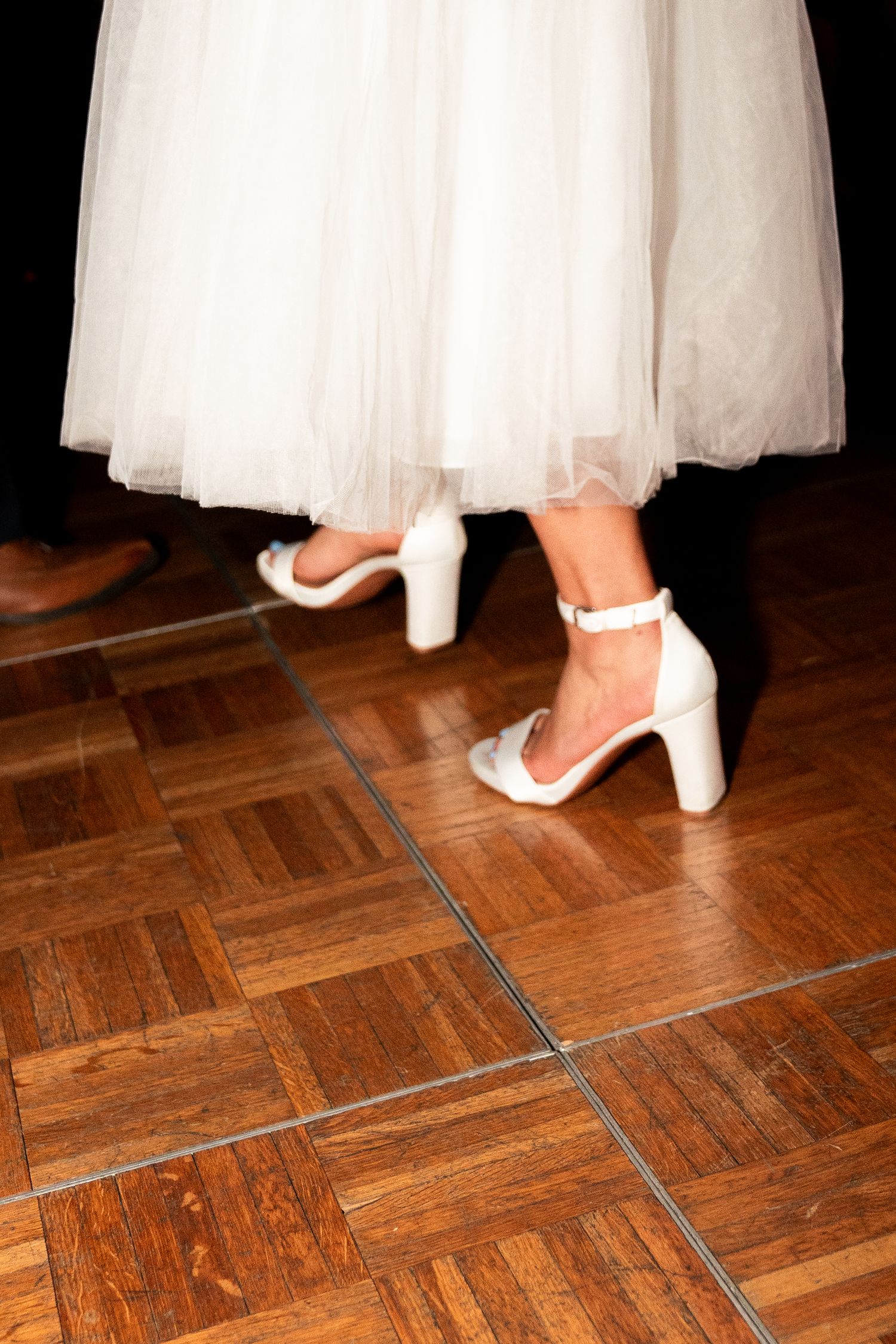 A close-up of white block heel sandals and tulle dress hem on a parquet dance floor.