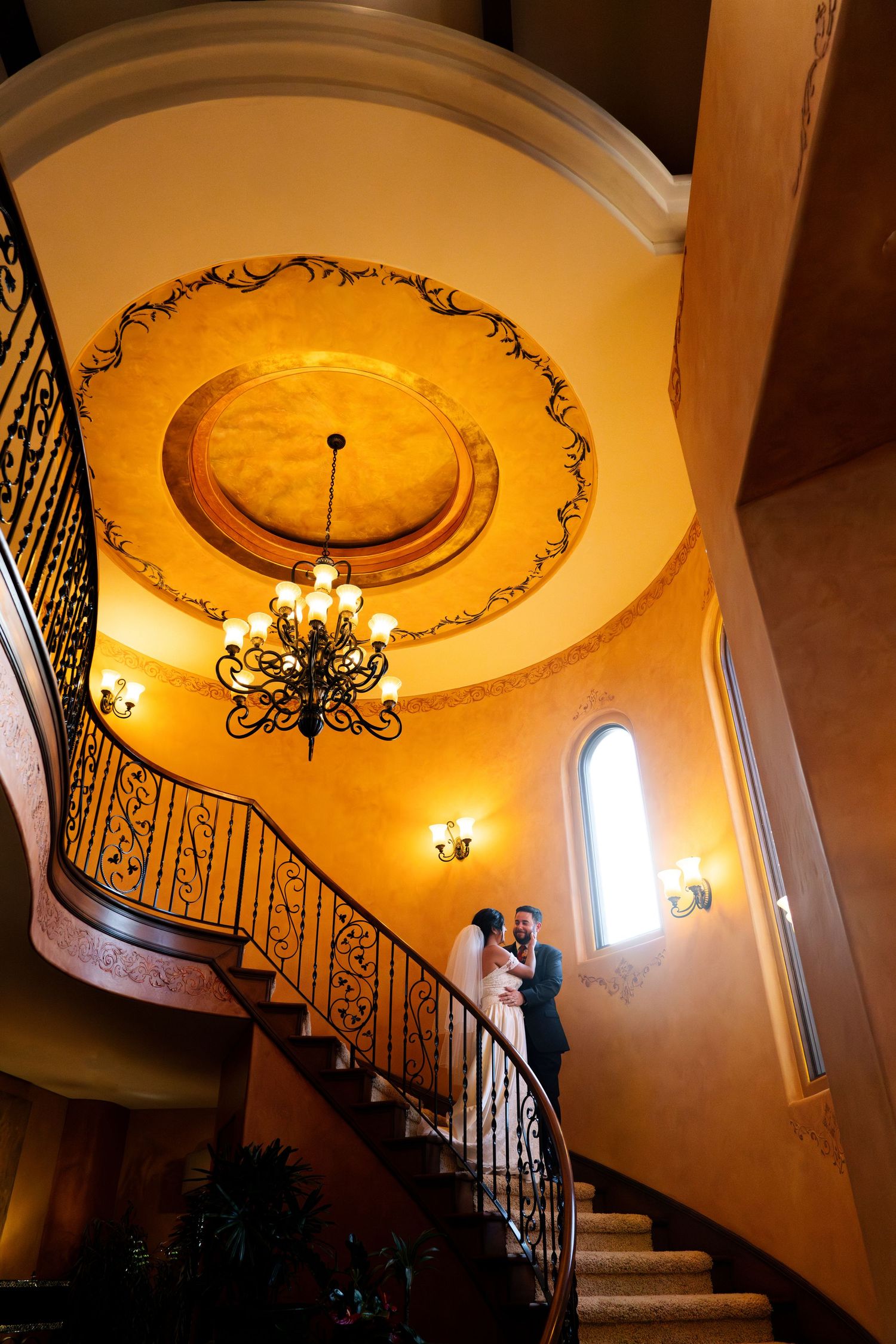 A romantic silhouette at a grand staircase under an ornate ceiling with chandelier in warm ambient lighting.