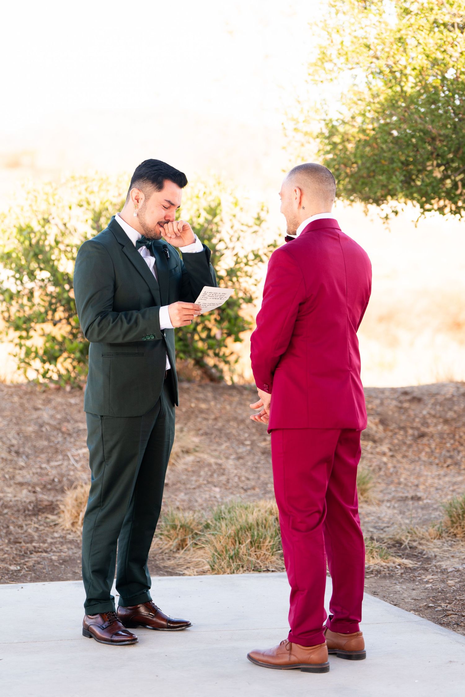 Emotional wedding moment between couple in dark green and burgundy suits sharing vows outdoors during ceremony.