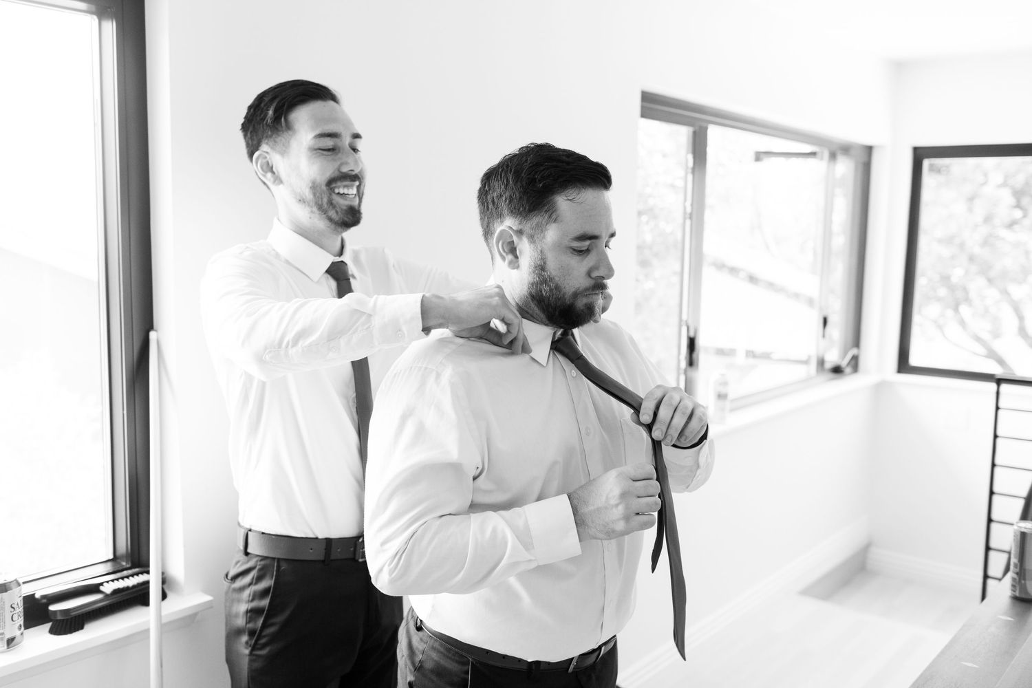 Two men in white dress shirts share a candid moment while adjusting a tie in a bright sunlit room.