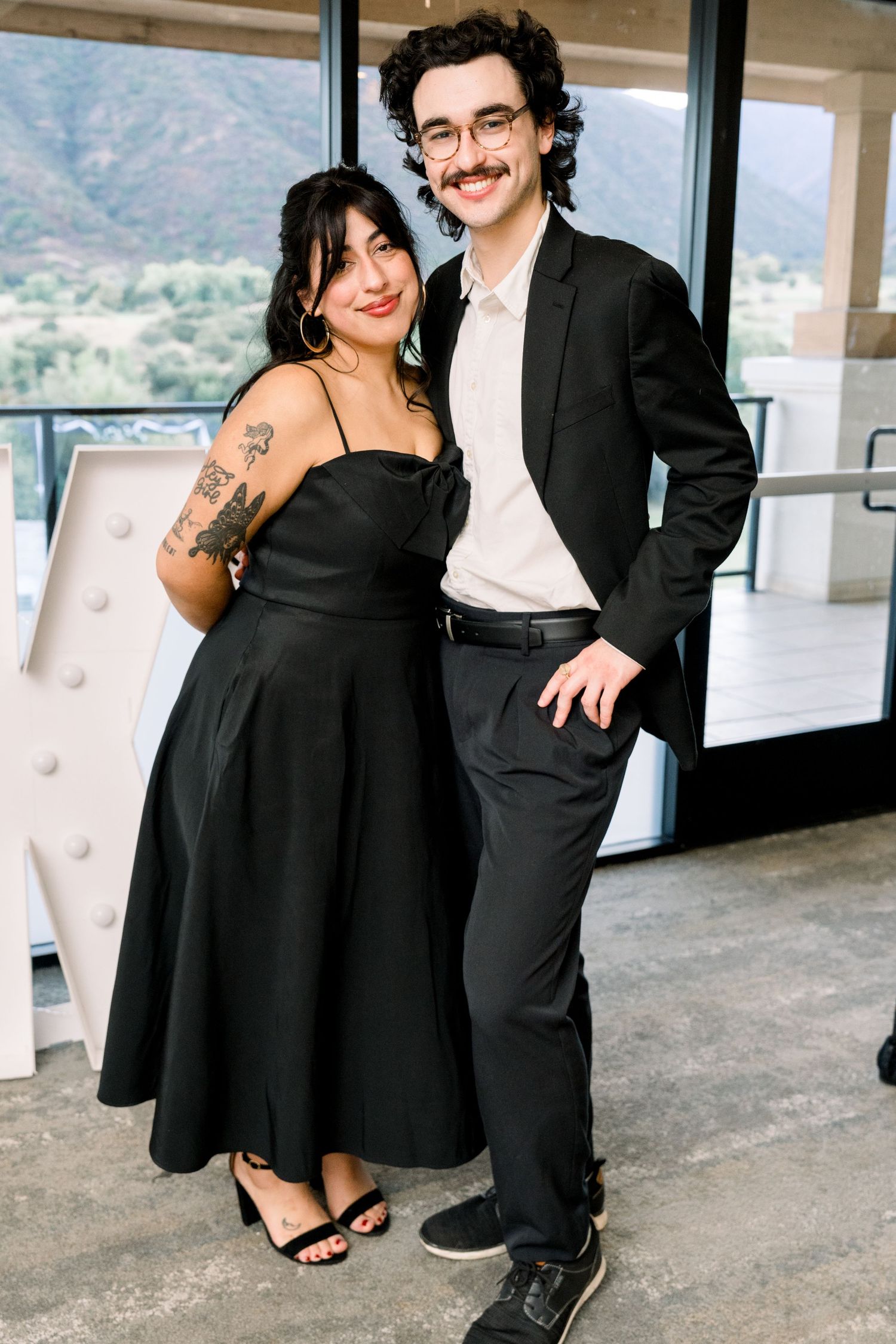 A couple in formal attire poses together on a balcony with mountain views in the background.