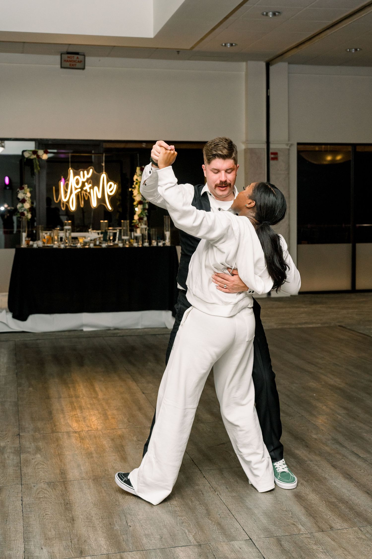Two people dancing together in formal attire against a dark backdrop with a bar table in the background.