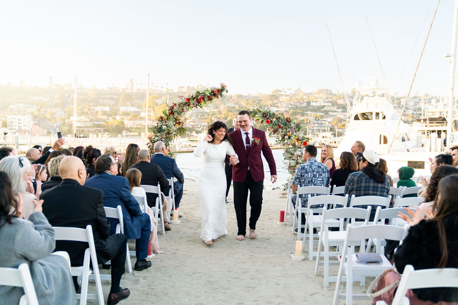 A couple walks down a beach wedding aisle with white chairs and guests on a sunny day.
