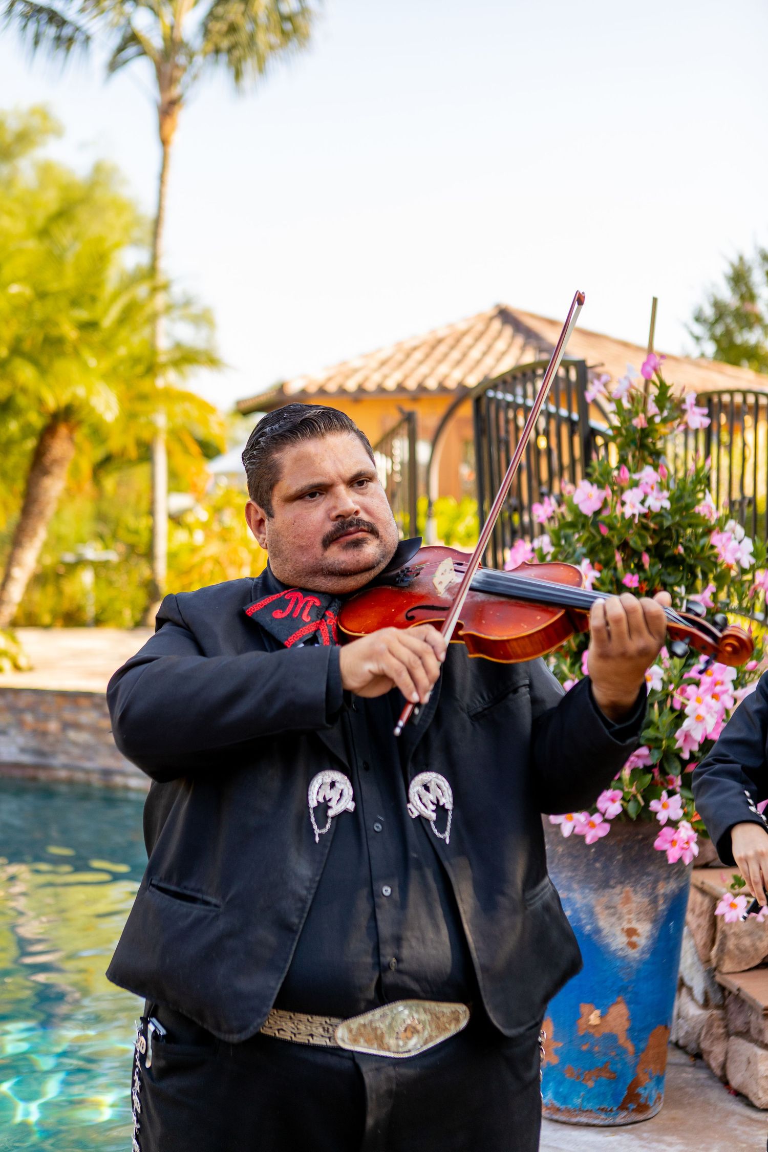 Mariachi violinist in traditional black suit performing by a pool with flowering garden.
