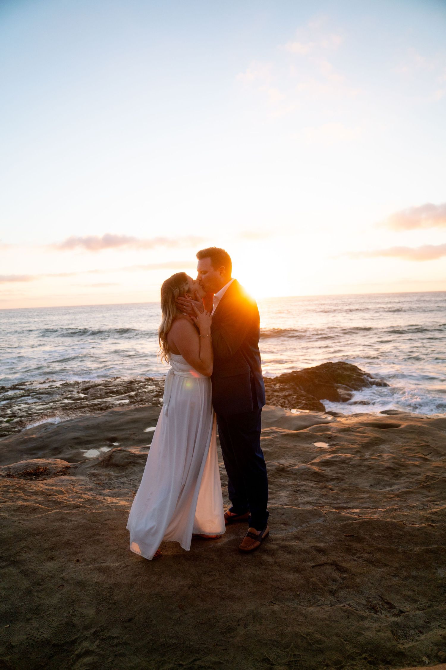 A couple shares a romantic kiss on rocky beach cliffs during a beautiful golden sunset over the ocean.