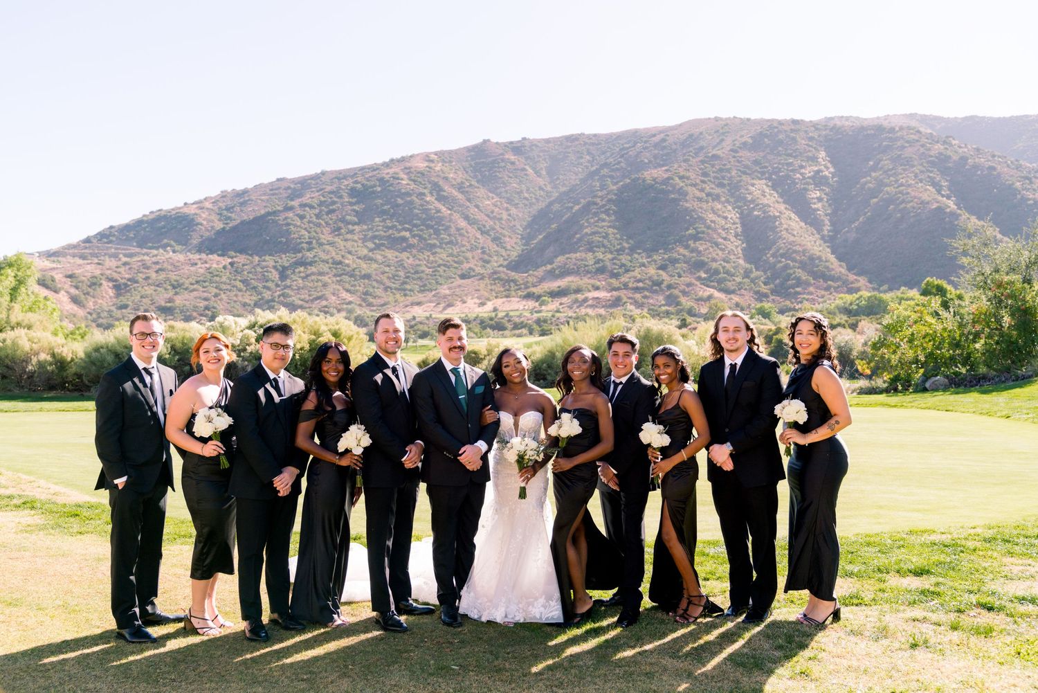 Wedding party in black attire poses together for group photo with mountain landscape in background.